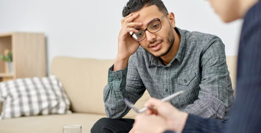 Man in glasses looking worried, sits attentively on a beige couch during a therapy session with a therapist opposite him.