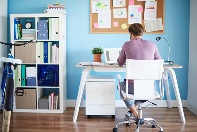Person sitting at white desk in organized home office with blue walls, bulletin board, and white storage shelving