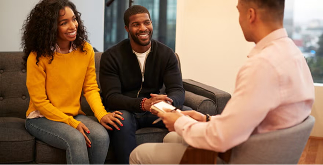 Smiling couple sitting on dark couch engaged in relationship counseling session with male therapist in bright office