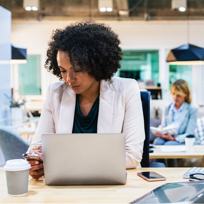 Woman with natural curly hair in white blazer focused on laptop work at desk with coffee in bright modern office