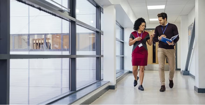 Two diverse professionals walking and discussing while reviewing documents in bright modern office hallway