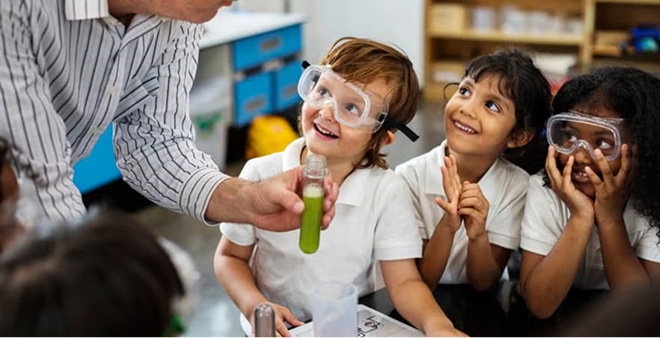 Teacher in striped shirt demonstrating science experiment with test tube to three diverse elementary students wearing safety goggles in classroom
