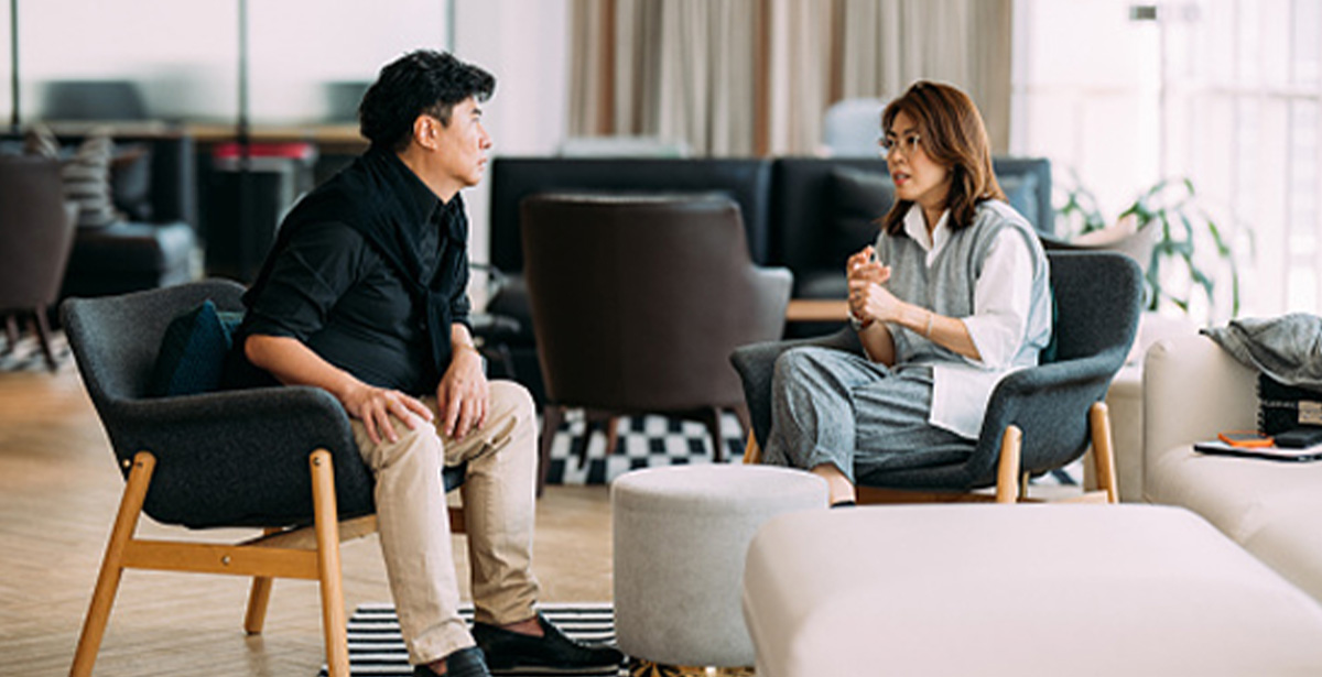 A man and woman sitting in office chairs, engaged in conversation in a professional office setting.