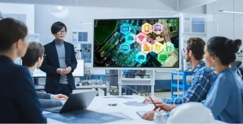 Project manager presenting technology infographic on large screen to diverse team of professionals seated at conference table in bright modern office