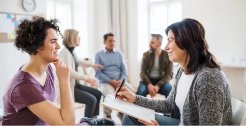 Counselor taking notes during group therapy session with clients conversing in background