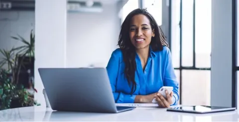Professional woman in blue blouse smiling at desk with laptop and coffee in bright modern office