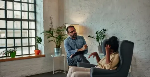 Psychologist in denim shirt sitting with clipboard talking to distressed client in grey armchair in bright therapy office with plants