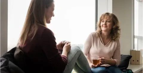 A parent and teenage child sitting together on a couch at home, talking closely and smiling gently, suggesting emotional support.