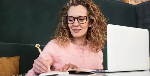 Professional woman writing in notebook while working on laptop at home