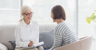 A counsellor is sitting on a couch opposite a client. She is taking notes and smiling.